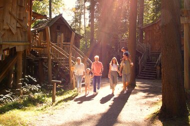 Familie spaziert bei warmem Licht durch das Natur-Resort Tripsdrill. Entlang ihres Weges stehen mehrere Baumhäuser aus Holz zwischen hohen Bäumen. | © Erlebnispark Tripsdrill