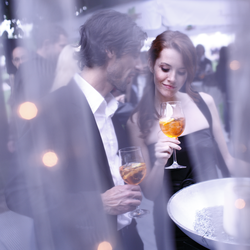 Ein elegantes Paar steht bei einer Veranstaltung im Freien, beide mit einem Glas Aperitif in der Hand. Die Frau, in einem schwarzen Abendkleid, betrachtet ihr Getränk mit einem sanften Lächeln, während der Mann in einem Anzug ihr nahe steht. Durch einen Schleier von Lichtern im Vordergrund wird eine romantische und stilvolle Atmosphäre geschaffen. Im Hintergrund sind unscharfe Silhouetten anderer Gäste und eine lebhafte Veranstaltung zu erkennen. | © TMBW | Christoph Düpper