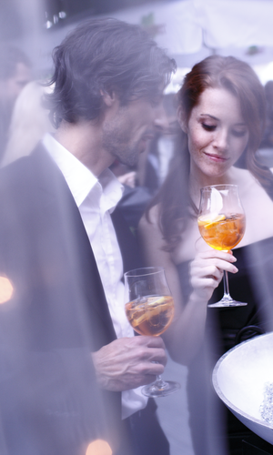 Ein elegantes Paar steht bei einer Veranstaltung im Freien, beide mit einem Glas Aperitif in der Hand. Die Frau, in einem schwarzen Abendkleid, betrachtet ihr Getränk mit einem sanften Lächeln, während der Mann in einem Anzug ihr nahe steht. Durch einen Schleier von Lichtern im Vordergrund wird eine romantische und stilvolle Atmosphäre geschaffen. Im Hintergrund sind unscharfe Silhouetten anderer Gäste und eine lebhafte Veranstaltung zu erkennen. | © TMBW | Christoph Düpper