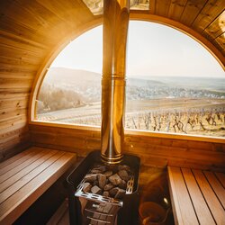 Im Inneren der Panoramasauna aus Holz sorgt ein glänzender Saunaofen für wohlige Wärme. Durch das große, bogenförmige Fenster eröffnet sich ein eindrucksvoller Blick auf die Rebhänge und die malerische Landschaft des Land der 1000 Hügel. | © Bofinger & König GbR