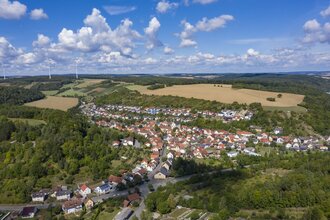 Luftbildaufnahme bei strahlendem Sonnenschein auf den Ort Dittwar. Der Ort liegt inmitten von Feldern und Wald. Im Hintergrund sind Windräder zu sehen. | © Tourist-Info Tauberbischofsheim