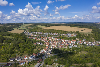 Luftbildaufnahme bei strahlendem Sonnenschein auf den Ort Dittwar. Der Ort liegt inmitten von Feldern und Wald. Im Hintergrund sind Windräder zu sehen. | © Tourist-Info Tauberbischofsheim