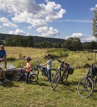 Zwei Erwachsene und zwei Kinder legen während einer Radtour eine Pause an der Tauber ein. Die Erwachsenen sitzen auf einer Holzbank. Der Bereich wurde renaturiert, Die Tauber ist im Hintergrund. zu sehen. Sie fließt im sanftem Bogen vorbei. | © Tourist-Info Tauberbischofsheim