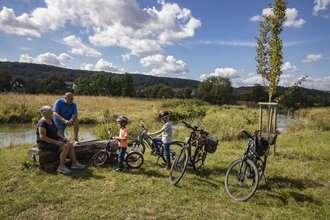 Zwei Erwachsene und zwei Kinder legen während einer Radtour eine Pause an der Tauber ein. Die Erwachsenen sitzen auf einer Holzbank. Der Bereich wurde renaturiert, Die Tauber ist im Hintergrund. zu sehen. Sie fließt im sanftem Bogen vorbei. | © Tourist-Info Tauberbischofsheim