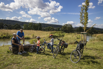 Zwei Erwachsene und zwei Kinder legen während einer Radtour eine Pause an der Tauber ein. Die Erwachsenen sitzen auf einer Holzbank. Der Bereich wurde renaturiert, Die Tauber ist im Hintergrund. zu sehen. Sie fließt im sanftem Bogen vorbei. | © Tourist-Info Tauberbischofsheim