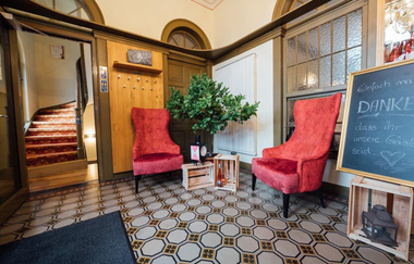 Im Eingangsbereich stehen zwei rote Sessel und eine Tafel, im Hintergrund sieht man eine geschwungene Treppe | © Touristikgemeinschaft Hohenlohe e. V.