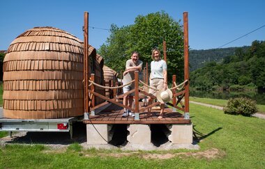 Es ist ein Holziglu auf einem Hänger zu sehen. Davon eine Holzterrasse, auf der 2 Frauen stehen.
