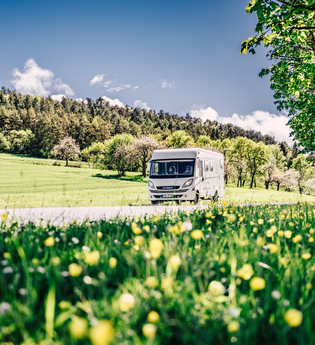 Weißes Wohnmobil fährt auf einer Strasse. Im Vordergrund ist eine Blumenwiese im Hintergrund eine Steuobstwiese mit angrenzendem Wald. | Odenwald | © Touristikgemeinschaft Odenwald e.V.