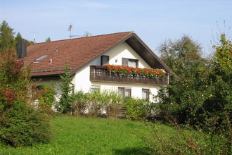 Haus mit rotem Satteldach und Balkon mit roten Blumen, umgeben von grünen Büschen und Wiese | © Ferienhof Schwab