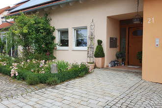 Eingangsbereich eines Hauses mit brauner Tür, Hausnummer 21, Blumenbeet mit Rosen und grünen Pflanzen neben gepflastertem Weg | © Familie Bokmeier