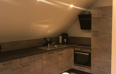 Moderne Küche unter einer Dachschräge mit grauen Schränken, Herd, Spüle und einem weißen Hochstuhl vor einem Tisch | © Brennerei Stilling