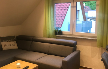 Wohnzimmer mit grauem Ecksofa, zwei Kissen mit der Aufschrift 'Auszeit', kleinem Holztisch mit Kerze und Fernbedienung, Fenster mit grünen Vorhängen und Blick auf Nachbarhäuser | © Brennerei Stilling