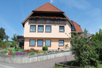 Zweistöckiges Ferienhaus mit roten Dachziegeln, Balkon mit Blumenkästen und gepflegtem Vorgarten unter blauem Himmel | © Ferienwohnung Ehrmann