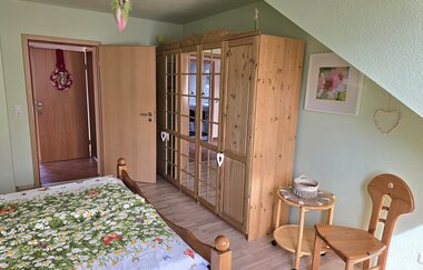 Schlafzimmer mit Holzbett, Blumendecke, großem Holzkleiderschrank mit Spiegeltüren, kleinem runden Beistelltisch und Holzstuhl unter einer Dachschräge | © Familie Fuhrmann