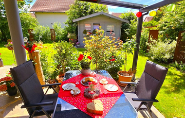 Gedeckter Gartentisch mit rotem Tischtuch, Kuchen und Tassen, umgeben von zwei gepolsterten Stühlen, im Hintergrund ein Gartenhaus und grüne Pflanzen | © Familie Fuhrmann