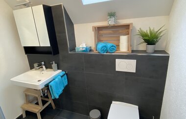 Modernes Badezimmer mit weißer Toilette, weißem Waschbecken auf Holzgestell, grauen Fliesen und Fenster mit Tageslicht | © Ferienwohnung Etzl