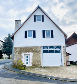 Weißes Haus mit steilem Dach, drei Fenstern mit schwarzen Fensterläden, einer weißen Tür und einer großen weißen Garagentür, im Hintergrund ein Glockenturm | © Ferienwohnung Etzl