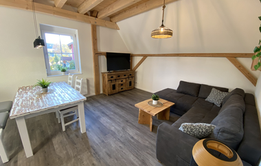 Wohn- und Esszimmer mit Holzbalkendecke, grauer Eckcouch, kleinem Holztisch, Esstisch mit weißen Stühlen und Fernseher auf Holzkommode | © Ferienwohnung Etzl