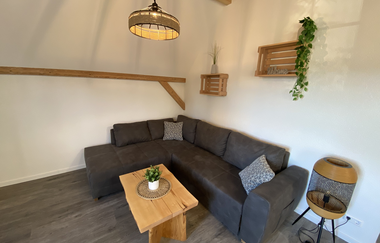 Wohnzimmer mit grauer Eckcouch, hellem Holztisch, hängender Lampe und Wandregalen mit Pflanzen und Dekoration | © Ferienwohnung Etzl
