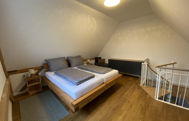 Schlafzimmer mit Doppelbett, grauer Bettwäsche, Holzboden, Nachttisch mit Lampe und Treppe mit Metallgeländer | © Ferienwohnung Etzl
