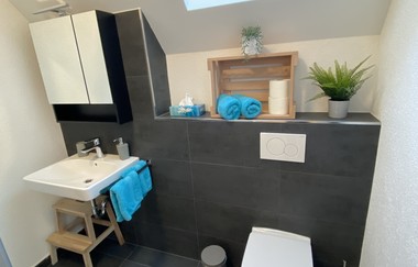 Modernes Badezimmer mit weißer Toilette, weißem Waschbecken auf Holzgestell, grauen Fliesen und Fenster mit Tageslicht | © Ferienwohnung Etzl