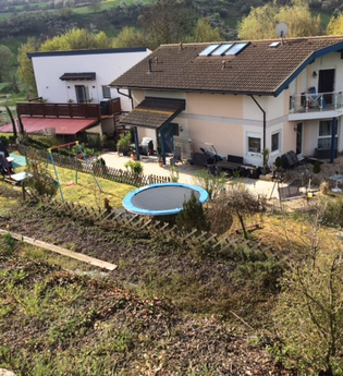 Zweistöckiges Haus mit Balkon und Terrasse, Garten mit Trampolin und Schaukel, umgeben von Bäumen und Hügeln im Hintergrund | © Jürgen Bauer