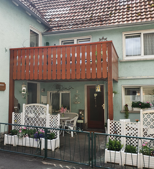 Eingang eines zweistöckigen Hauses mit grünem Putz, braunem Balkon und weißem Gartenzaun mit Blumenkästen davor | © Irene Peppel