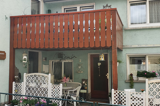 Eingang eines zweistöckigen Hauses mit grünem Putz, braunem Balkon und weißem Gartenzaun mit Blumenkästen davor | © Irene Peppel