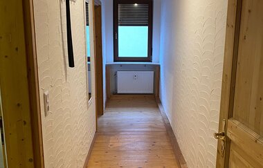 Schmaler Flur mit Holzboden, weißen Wänden und Holzdecke, Fenster am Ende mit geschlossener Jalousie, Garderobenhaken und Spiegel an der linken Wand | © Ferienwohnung Etzl