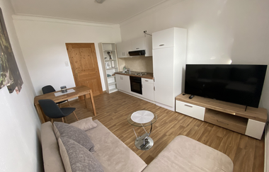 Kleines Wohn-Esszimmer mit offener Küche, beigem Sofa, Glastisch, Esstisch mit zwei Stühlen, TV auf Holz-TV-Board und Holztür | © Ferienwohnung Etzl