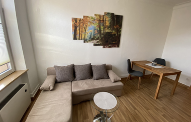 Wohnzimmer mit beiger Eckcouch, drei grauen Kissen, rundem Glastisch, Holztisch und zwei schwarzen Stühlen, Wandbild mit Waldmotiv | © Ferienwohnung Etzl