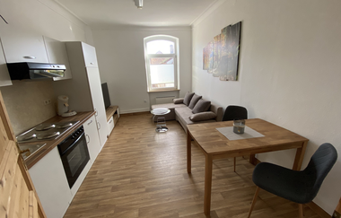 Kleines Wohn- und Esszimmer mit Küchenzeile, Holzboden, grauem Sofa, Holztisch mit zwei Stühlen und Fenster mit halb geschlossener Jalousie | © Ferienwohnung Etzl