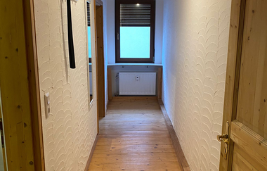 Schmaler Flur mit Holzboden, weißen Wänden und Holzdecke, Fenster am Ende mit geschlossener Jalousie, Garderobenhaken und Spiegel an der linken Wand | © Ferienwohnung Etzl