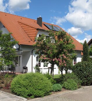 Weißes Einfamilienhaus mit rotem Ziegeldach und Solarpanelen, umgeben von grünen Büschen und blühendem Baum, bei sonnigem Himmel | © Erika Zeller