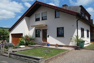 Einfamilienhaus mit weißer Fassade, braunem Garagentor, gepflegtem Vorgarten mit Blumen und Palmen, unter blauem Himmel | © Familie Noe
