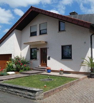 Einfamilienhaus mit weißer Fassade, braunem Garagentor, gepflegtem Vorgarten mit Blumen und Palmen, unter blauem Himmel | © Familie Noe