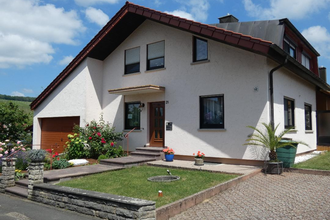 Einfamilienhaus mit weißer Fassade, braunem Garagentor, gepflegtem Vorgarten mit Blumen und Palmen, unter blauem Himmel | © Familie Noe