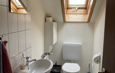 Kleines Badezimmer mit Toilette unter einem Dachfenster, Waschbecken, Spiegel und blauer Badvorleger mit Meeresmotiven