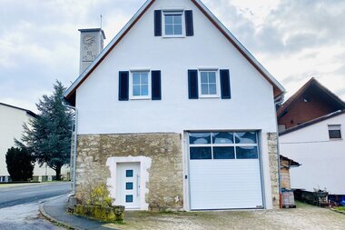 Zweistöckiges Haus mit weißer Fassade und steilem Dach, vier Fenstern mit schwarzen Fensterläden, weißer Haustür und großer Garagentür, im Hintergrund ein Glockenturm mit Uhr | © Ferienwohnung Etzl