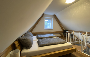 Schlafzimmer unter Dachschräge mit Doppelbett, grauen Bettdecken, Fenster und Treppe mit Geländer | © Ferienwohnung Etzl