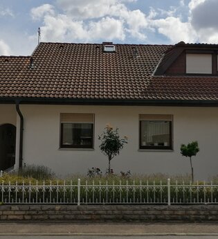 Einfamilienhaus mit weißer Fassade, braunem Ziegeldach, zwei Fenstern, Garage und kleinem Garten mit Büschen und Bäumen | © Ferienwohnung Gschwind