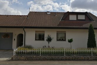 Einfamilienhaus mit weißer Fassade, braunem Ziegeldach, zwei Fenstern, Garage und kleinem Garten mit Büschen und Bäumen | © Ferienwohnung Gschwind