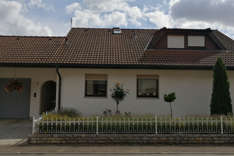 Einfamilienhaus mit weißer Fassade, braunem Ziegeldach, zwei Fenstern, Garage und kleinem Garten mit Büschen und Bäumen | © Ferienwohnung Gschwind
