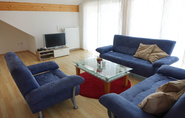 Wohnzimmer bestehend aus drei bequemen blauen Couchen und einem Tisch in der Mitte, an der Wand steht ein Fernseher und es gibt eine große Fensterfront | © Neckar-Zaber-Tourismus e.V.