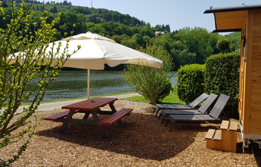 Ein Fluss dahienr eine Sitzgruppe aus Hol mit einem weissen Sonnenschrift. Dahinter stehen zwei Sonnenliegen mit einem Schäferwagen. Binau im Odenwald | © Fortuna Camping Binau
