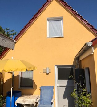 Gelbes Haus mit rotem Dach, weißer Tür, grauer Garage und Terrasse mit blauem Stuhl, gelbem Sonnenschirm und kleinem Tisch | © Gästehaus Sabine