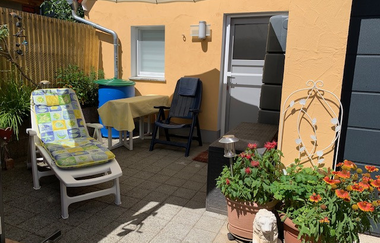 Sonnige Terrasse mit Liegestuhl, Tisch mit gelber Tischdecke, zwei Stühlen, großen Blumentöpfen mit roten und orangen Blumen und einem weißen Sonnenschirm | © Gästehaus Sabine