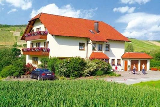 Zweistöckiges Haus mit rotem Dach, Balkonen mit Blumen, umgeben von grünen Feldern und zwei Personen mit Fahrrad vor dem Eingang | © Gästehaus Schieser