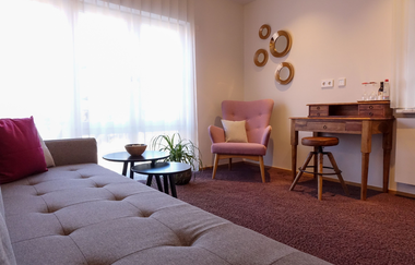 Aufnahme eines Zimmers mit Wohnbereich: graue Couch, zwei kleine schwarze Tische, ein rosa Sesselstuhl und ein hölzerner Schreitisch. Roter Teppichboden. | © Neckar-Zaber-Tourismus e.V.
