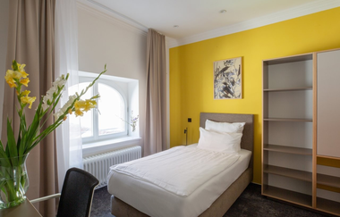 Hotelzimmer mit Einzelbett und kleinem Tisch mit Stuhl. Auf dem Tisch befindet sich eine Blumenvase mit gelben Blumen, | © Hotel Kredell